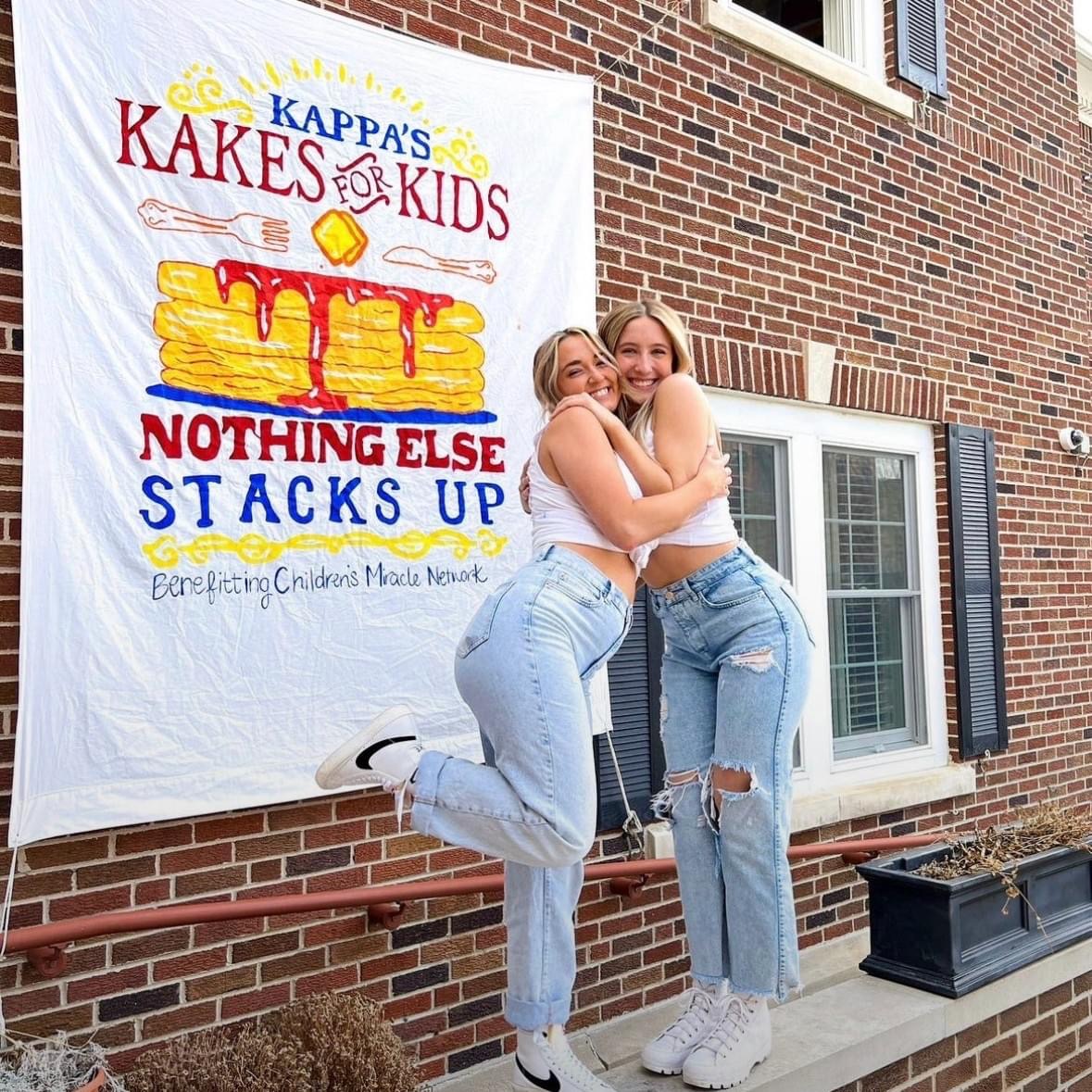 photo of two women hugging in front a banner that reads Kappas Kakes for Kids